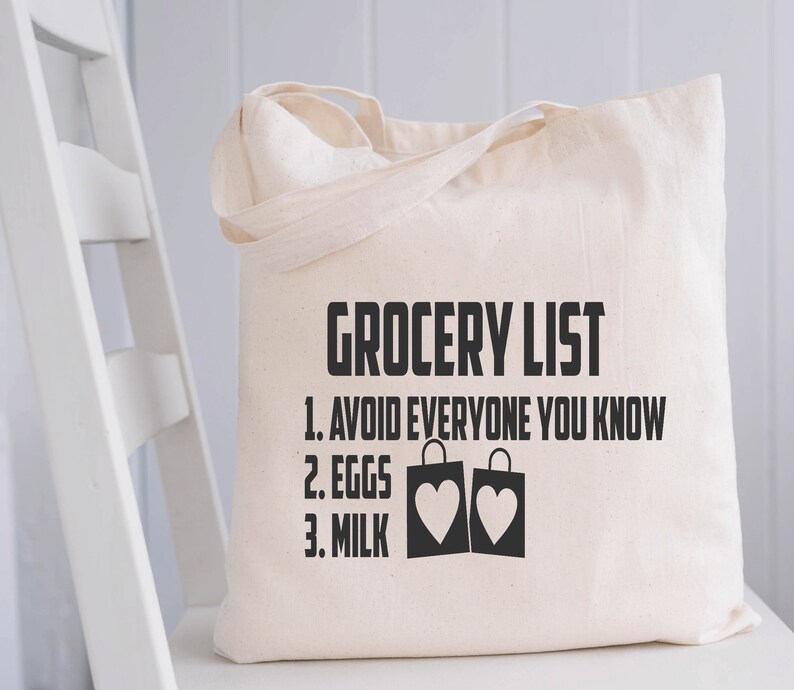 May include: Cream-colored canvas tote bag with black text that reads "GROCERY LIST" followed by a list: "1. AVOID EVERYONE YOU KNOW", "2. EGGS", and "3. MILK". The bag features two shopping bags with heart shapes.