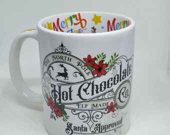 Może przedstawiać: Biały kubek ceramiczny z czerwono-zielono-czarnym wzorem z napisem "The North Pole Hot Chocolate Co. Elf Made Santa Approved". Wewnątrz kubka znajduje się kolorowy wzór "Merry".