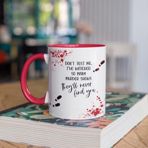 May include: White ceramic mug with a red rim and handle. The mug has a red splatter pattern and footprints. The text on the mug reads "Don't test me. I've watched so many murder shows they'll never find you."