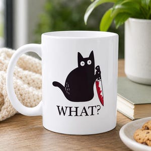 May include: A white ceramic mug featuring a black cat illustration holding a knife with red blood stains on the blade. Below the cat, the word WHAT? is printed in black capital letters. The mug has a curved handle and a smooth, glossy finish.