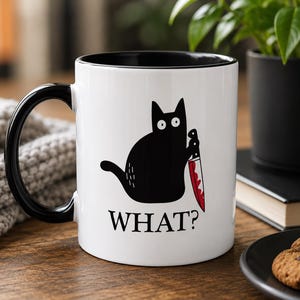 May include: A white ceramic mug with a black handle and interior. The front features a graphic of a black cat with wide eyes holding a knife with red blood stains on the blade. Below the cat, the word WHAT? is printed in black capital letters.