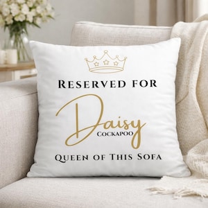 May include: White square throw pillow with gold crown graphic and text that reads "RESERVED FOR Daisy COCKAPOO QUEEN OF THIS SOFA" in black and gold lettering. The pillow is on a beige sofa.