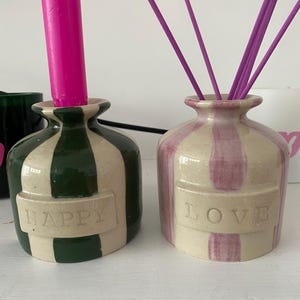 May include: Two ceramic vases. One is green and white striped with a pink candle, the other pink and white striped with purple reeds. The green vase says "HAPPY", the pink vase says "LOVE".
