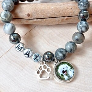 May include: Gray and black beaded bracelet with a silver charm of a paw print and a round charm with a photo of a white dog. The bracelet has silver beads with the letters "MAX" on them.