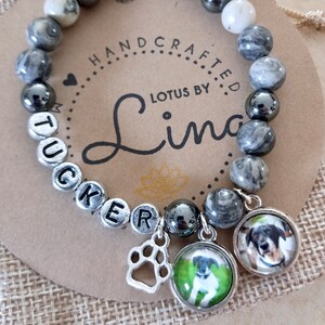 May include: A gray and white marble bracelet with silver beads spelling out the name 'TUCKER'. The bracelet has two charms, one is a silver paw print and the other is a glass circle with a picture of a dog inside. The bracelet is on a brown background with the words 'HANDCRAFTED LOTUS BY Lina' printed on it.