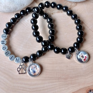 May include: Two black beaded bracelets. One bracelet has the name "THORIN" spelled out in silver letter beads. Both bracelets have a charm with a dog's photo and a paw print charm. The bracelets are on a wooden surface.