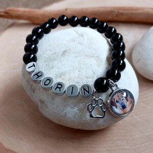 May include: Black beaded bracelet with silver beads spelling "Thorin" and a charm with a paw print and a photo of a dog.