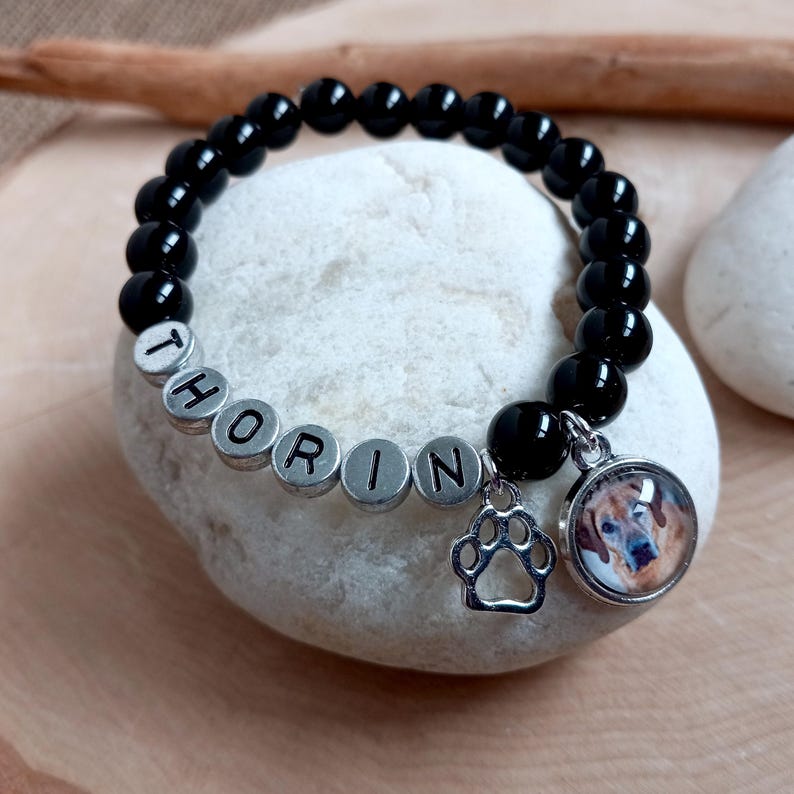 May include: Black beaded bracelet with silver beads spelling "Thorin" and a charm with a paw print and a photo of a dog.