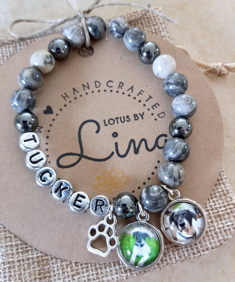 May include: A gray and white marble bracelet with silver beads spelling out the name 'TUCKER'. The bracelet has two charms, one is a silver paw print and the other is a glass circle with a picture of a dog inside. The bracelet is on a brown background with the words 'HANDCRAFTED LOTUS BY Lina' printed on it.