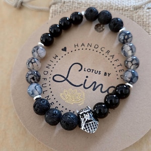 May include: A beaded bracelet featuring black onyx, lava rock, and patterned gray beads. The bracelet includes a silver-colored charm and is displayed on a card with the text "Lotus by Lina".