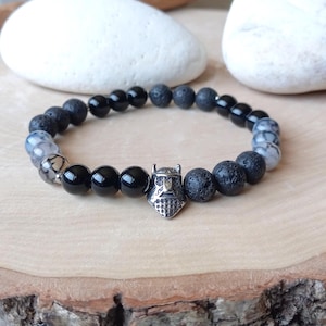May include: A beaded bracelet featuring black onyx, lava rock, and patterned stone beads. A silver-colored charm in the center depicts a Viking helmet. The bracelet is strung on an elastic band.