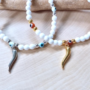 May include: White beaded bracelets with silver and gold accents. Each bracelet features a charm, one silver horn and one gold horn. The bracelets also have small evil eye beads in blue and red.