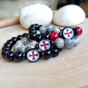May include: Two black and grey beaded bracelets with red and white cross charms. The bracelets are on a wooden surface.