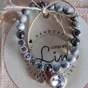 May include: Gray and black beaded bracelet with a silver charm of a paw print and a round charm with a picture of a dog. The bracelet has the name "BRONX" spelled out in silver beads. The tag attached to the bracelet reads "HANDCRAFTED LOTUS BY Lin JEWELRY".