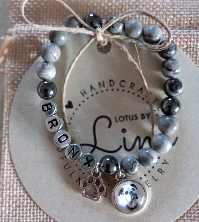 May include: Gray and black beaded bracelet with a silver charm of a paw print and a round charm with a picture of a dog. The bracelet has the name "BRONX" spelled out in silver beads. The tag attached to the bracelet reads "HANDCRAFTED LOTUS BY Lin JEWELRY".