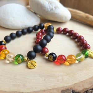 May include: Two beaded bracelets. One features matte black beads and colorful accent beads in red, yellow, green, and clear. The other bracelet has maroon beads and colorful accent beads. Both have gold charms.