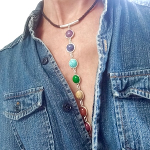 May include: A long leather necklace with a silver bar pendant and seven round gemstone beads in various colors. The necklace is worn by a person wearing a denim shirt.