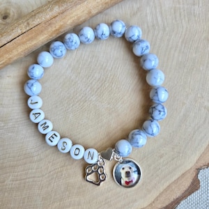 May include: A beaded bracelet with white and gray marbled beads. The bracelet includes white letter beads spelling "JAMESON", a paw print charm, and a circular charm with a dog's photo. The bracelet is on a wooden surface.