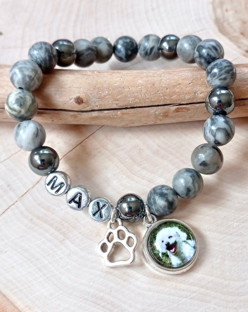 May include: Gray and black beaded bracelet with a silver charm of a paw print and a round charm with a photo of a white dog. The bracelet has silver beads with the letters "MAX" on them.