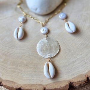 May include: A gold-toned necklace featuring a large, iridescent, circular pendant and several dangling elements. The necklace incorporates white seashells and pearls, creating a beachy, bohemian aesthetic. The necklace is displayed on a wooden surface.