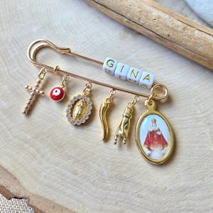 May include: Gold-toned safety pin with multiple charms, including a cross, an evil eye, a religious pendant, a horn, a hand gesture, and an oval pendant with a religious image. The name "GINA" is spelled out in white letter beads.