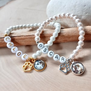 May include: Two white pearl bracelets with gold and silver paw charms. The bracelets have the names "Chloe" and "Oscar" spelled out in white letters on the pearls.