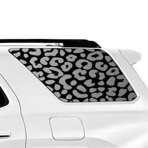 May include: A white SUV with a rear window featuring a black and grey leopard print design. The pattern consists of irregular, rounded shapes, creating a bold and stylish aesthetic. The vehicle's body is a bright white.