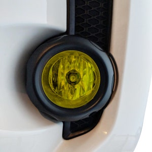 Könnte beinhalten: Nahaufnahme eines gelben Nebelscheinwerfers an einem weißen Auto. Der Nebelscheinwerfer ist rund und hat ein schwarzes Gehäuse.
