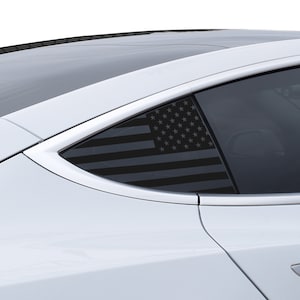May include: A black and gray American flag decal applied to the rear passenger window of a white car. The flag design is visible through the tinted glass, with the stars and stripes clearly defined. The car's body is white, and the window frame is silver.