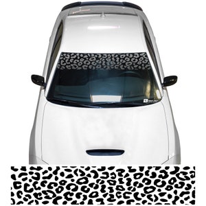 Puede incluir: Un coche blanco con un parasol de estampado de leopardo blanco y negro en el parabrisas. El parasol es una franja horizontal en la parte superior del parabrisas.
