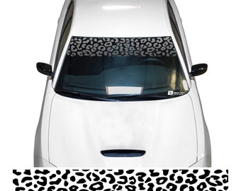 May include: A white car with a black and white leopard print sunshade on the windshield. The sunshade is a horizontal strip across the top of the windshield.