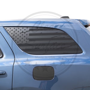 Può includere: Un design a bandiera americana nera è applicato su un finestrino dell'auto. Le stelle e le strisce della bandiera sono visibili, con un aspetto usurato e invecchiato. L'auto è di colore blu chiaro e il testo "Made in USA" è visibile.