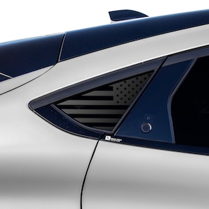 May include: A silver car with a black American flag decal on the rear window. The decal is in the shape of a triangle and is placed on the back window of the car.