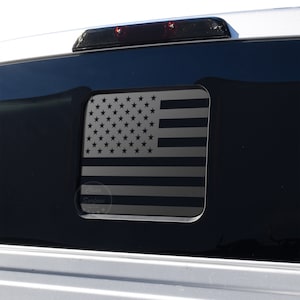May include: A black and white American flag decal on a black truck window. The flag is in a rectangular shape and has a white star field and red and white stripes.