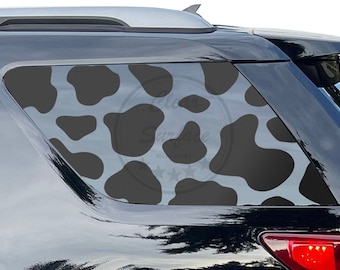 Op de afbeelding: Zwart-witte koe-print auto raam sticker. De sticker is aangebracht op de achterruit van een zwarte SUV.