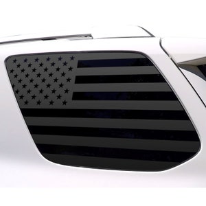 Könnte beinhalten: Schwarzer amerikanischer Flaggen-Fensteraufkleber, der auf einer Autoscheibe angebracht ist. Das Flaggen-Design zeigt Sterne und Streifen und bietet eine patriotische Ästhetik. Der Aufkleber hat ein dunkles, mattes Finish, das mit der weißen Karosserie kontrastiert.