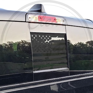 May include: A black truck with a rear window that has a white American flag decal. The flag is in the shape of the window and has a black background.
