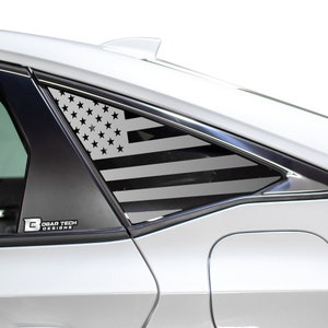 Puede incluir: Una calcomanía de bandera estadounidense en blanco y negro en la ventana de un automóvil. La calcomanía está en la ventana trasera del pasajero de un automóvil plateado. La calcomanía está hecha por Bogar Tech Designs.