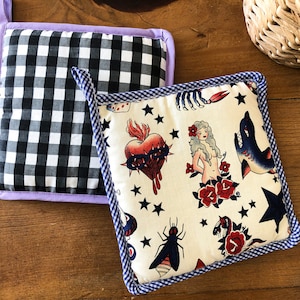 Pode incluir: Dois pegadores de panela, um com um padrão xadrez preto e branco e uma borda roxa, e o outro com um fundo branco e uma borda azul com um padrão repetido de tatuagens coloridas, incluindo um coração, uma mulher, um tubarão, uma abelha e uma cobra.