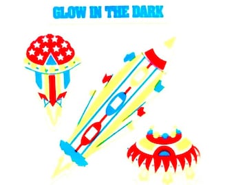 May include: Three glow-in-the-dark stickers featuring a rocket, a UFO, and a flying saucer. The rocket is yellow with blue and red accents. The UFOs are red, white, and blue.