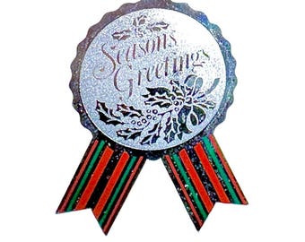 May include: A circular badge with a scalloped edge featuring a silver glitter finish and the text Seasons Greetings in dark script. Below the text is a black holly leaf and berry illustration. Two ribbons with red, green, and black stripes hang down.