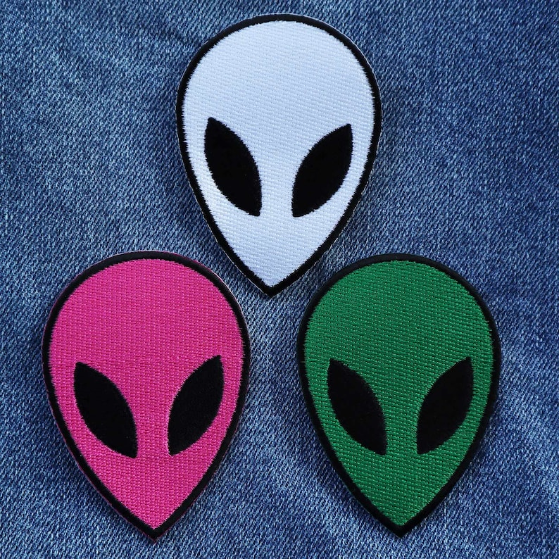 May include: Three alien head patches in different colours: white, pink and green. Each patch has black eyes and a simple alien head shape.