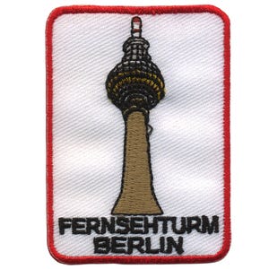 Könnte beinhalten: Ein weißer gestickter Aufnäher mit rotem Rand. Der Aufnäher zeigt eine braune und schwarze Illustration des Fernsehturms, eines Fernsehturms in Berlin, Deutschland. Der Text "Fernsehturm Berlin" ist in Schwarz unter dem Turm gestickt.