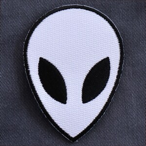 May include: White alien head patch with black eyes. The patch is made of fabric and has a stitched outline.