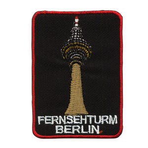 Puede incluir: Un parche bordado negro con un borde rojo. El parche presenta una imagen estilizada de la Fernsehturm Berlin, una torre de televisión en Berlín, Alemania. El texto "Fernsehturm Berlin" está bordado en blanco debajo de la torre.