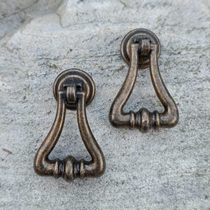 May include: Two antique bronze-toned drawer pulls. Each pull features a circular backplate and a triangular handle with decorative detailing. The pulls are made of metal and have a vintage aesthetic, suitable for furniture restoration or adding a classic touch.