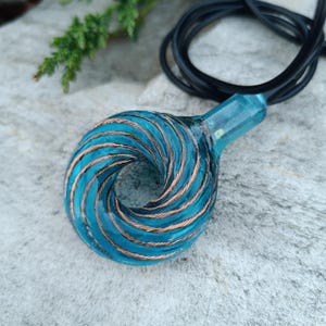 Può includere: Un ciondolo turchese con dettagli in filo di rame, sospeso a un cordino nero. Il ciondolo presenta un design a spirale. Lo sfondo è una superficie di pietra grigio chiaro.
