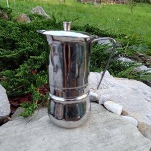 May include: A polished stainless steel stovetop espresso maker. The coffee pot has a rounded base, a cylindrical body, and a handle. The lid has a small, rectangular knob. The espresso maker is on a grey stone surface, with green foliage behind it.