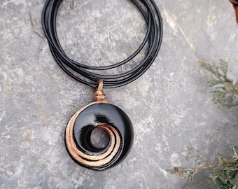 May include: A black pendant necklace with a spiral design. The pendant is a black circle with a copper-colored spiral in the center. The necklace has a black cord. The pendant is resting on a gray stone surface with green foliage.