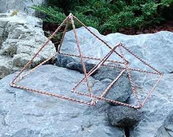 May include: Two copper wire pyramids, one larger than the other, rest on gray rocks. The pyramids are constructed with twisted wire, creating a geometric design. Green foliage is visible in the background.